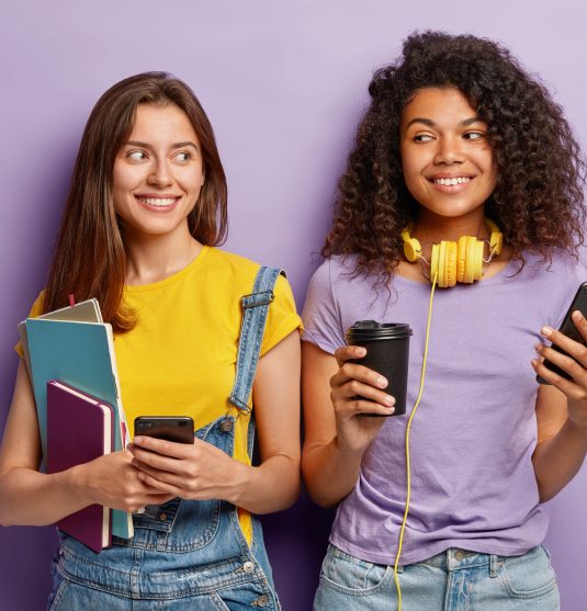 Indoor shot of pleasant looking cheerful two mixed race groupmates have coffee break after lectures, use modern cell phones for online communication listen radio in headphones carry notepad pocketbook