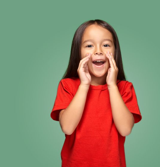 Portrait of a small pretty girl standing sideways and calling someone holding her hand near her mouth wearing a red t-shirt, green background